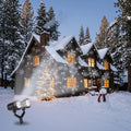 Dual-head snowflake projector in the snow projecting white snowflakes onto a wooden cabin at night, with a Christmas tree and snowman in the yard
