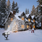 Dual-head snowflake projector in the snow projecting white snowflakes onto a wooden cabin at night, with a Christmas tree and snowman in the yard